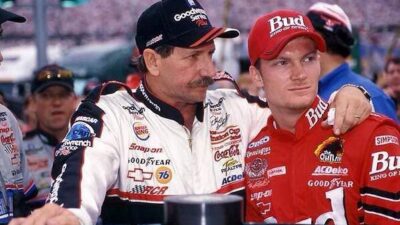 Two race car drivers in branded uniforms stand together at a race track event, showcasing camaraderie before a race.