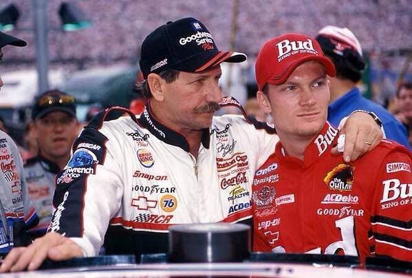 Two race car drivers in branded uniforms stand together at a race track event, showcasing camaraderie before a race.