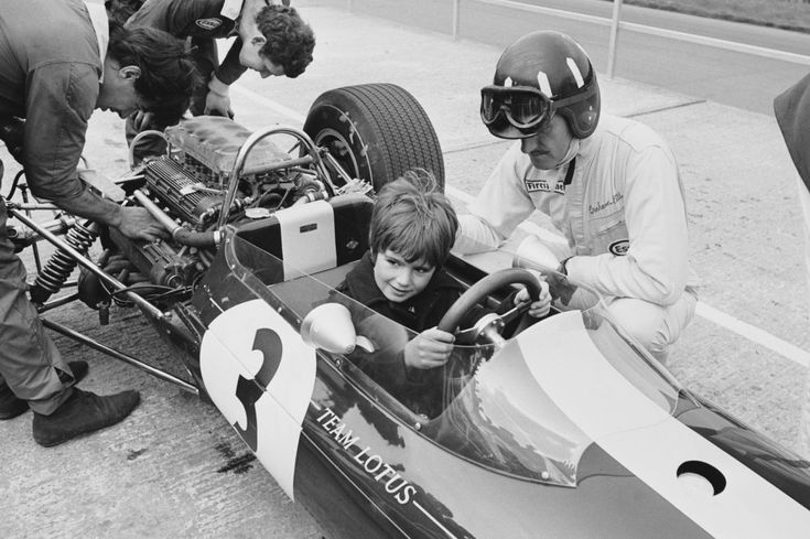 Child sits in Lotus race car with driver and mechanics nearby, 1960s racing scene, vintage motorsport moment.