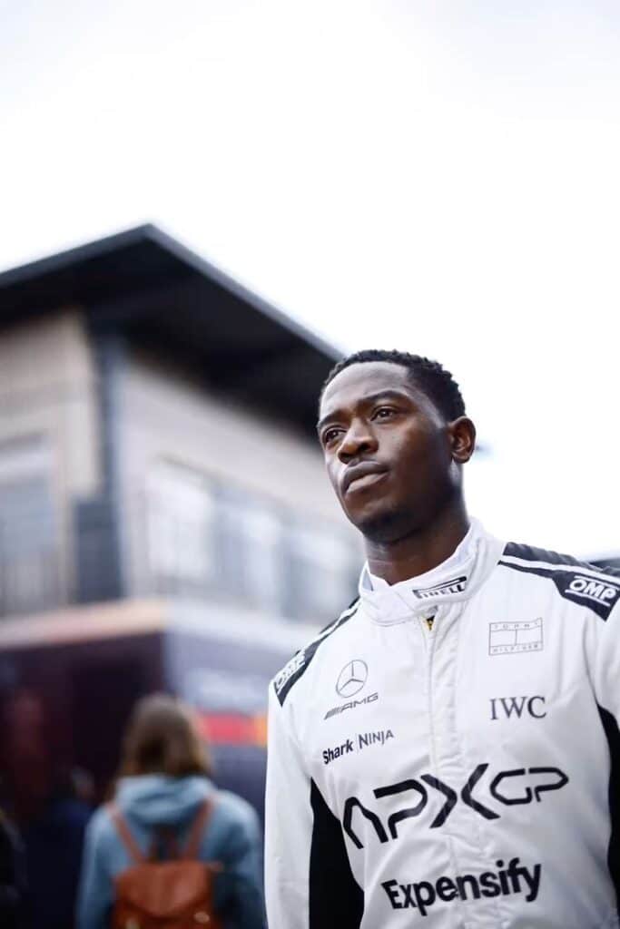 Race car driver in white suit with sponsor logos, standing outdoors, with a blurred building background.