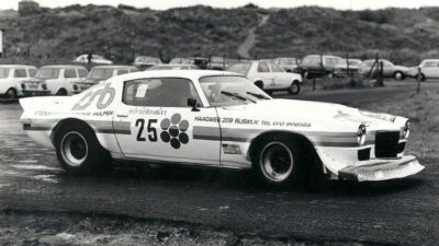 Vintage race car with number 25 on a wet track, surrounded by parked cars and hilly landscape in the background.