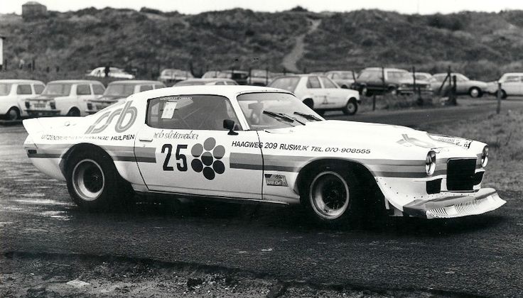 Vintage race car with number 25 on a wet track, surrounded by parked cars and hilly landscape in the background.