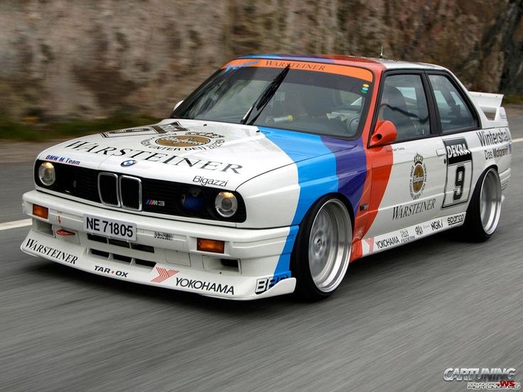 Vintage BMW E30 M3 race car speeding on a road, showcasing Warsteiner sponsorship and vibrant color livery.