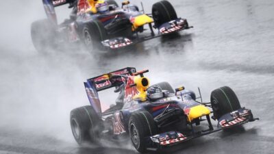 Two Formula 1 cars racing on a wet track, creating water spray behind them. Dynamic action shot capturing speed and precision.