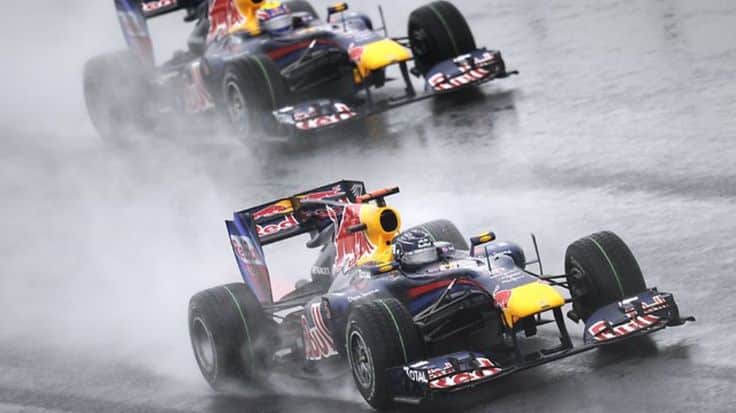 Two Formula 1 cars racing on a wet track, creating water spray behind them. Dynamic action shot capturing speed and precision.