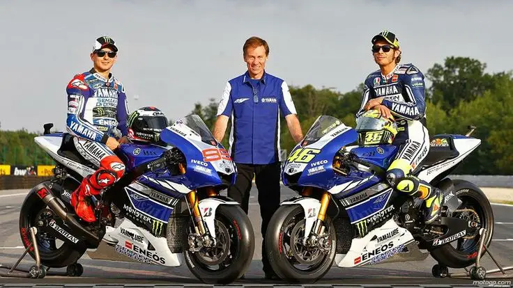 Two motorcyclists in Yamaha racing suits with bikes on a track, accompanied by a team member in a blue vest, ready for race.