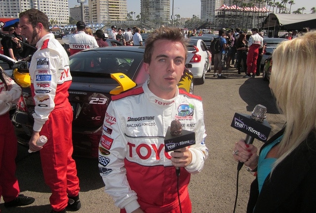 Race car driver being interviewed at a motorsport event, standing near a Toyota race car in a bustling paddock.