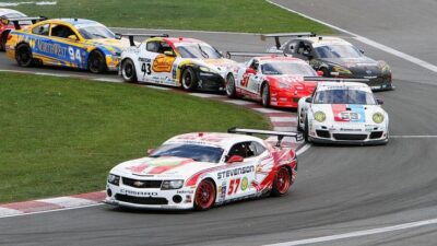 Race cars take a sharp turn in a competitive motorsport event, showcasing speed and precision on the asphalt track.