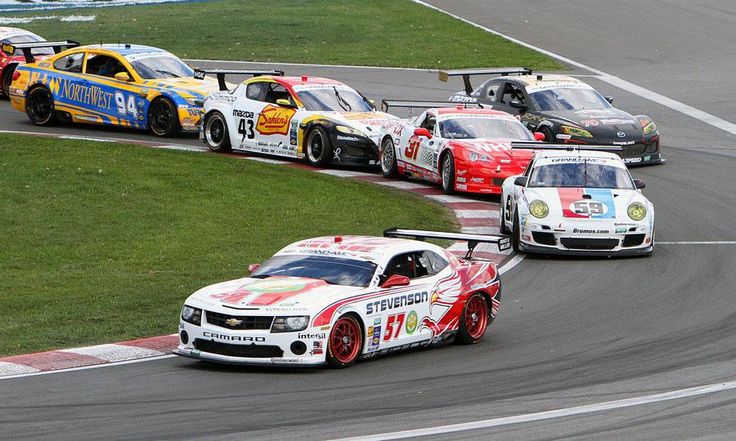 Race cars take a sharp turn in a competitive motorsport event, showcasing speed and precision on the asphalt track.