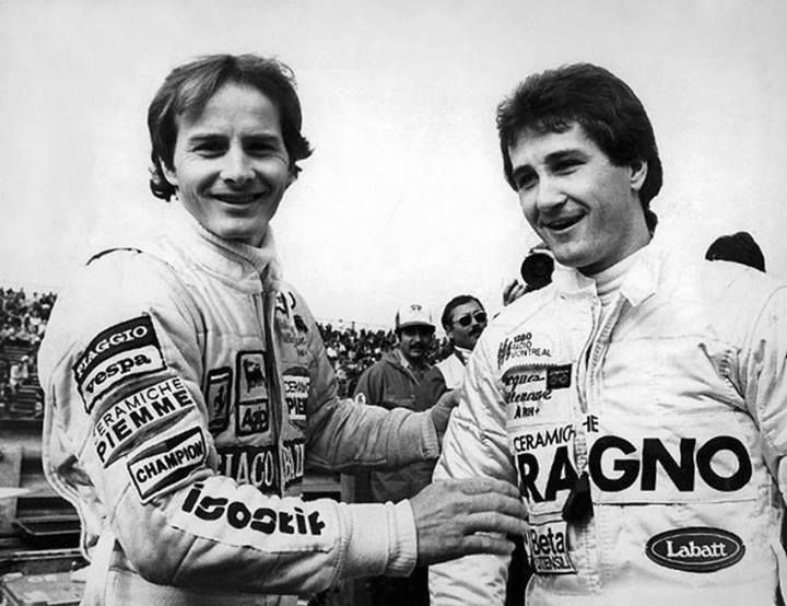 Two race car drivers in branded suits share a moment, smiling and interacting before a race event, with a crowd in the background.
