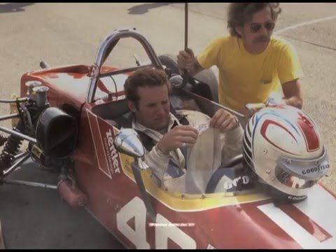 Race car driver prepares in cockpit, assisted by a man in yellow shirt, with helmet on side; vintage motorsport scene.