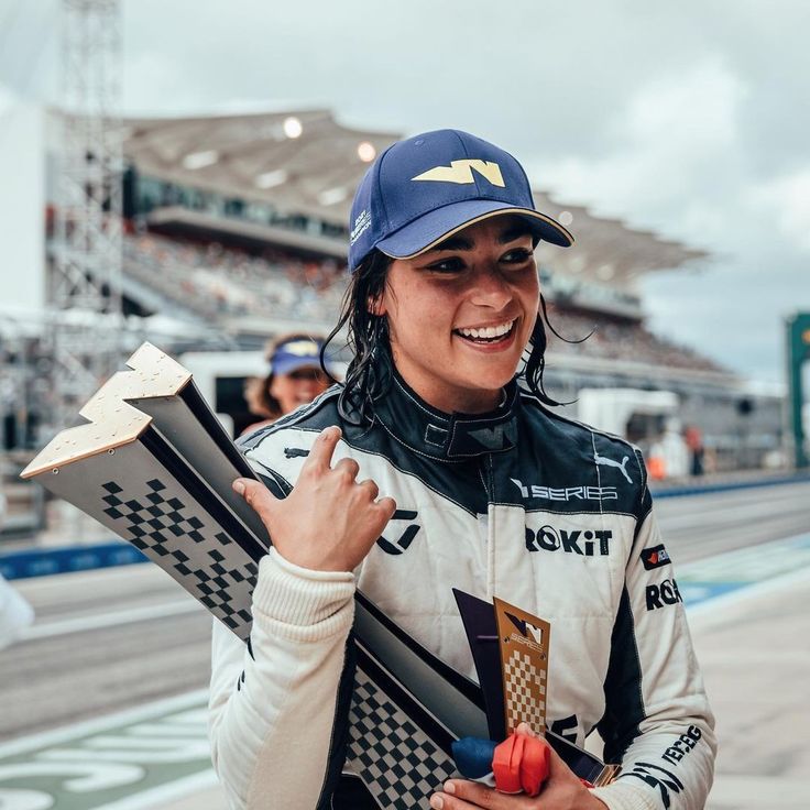 Race car driver celebrating with trophy at track, wearing a cap and racing suit.