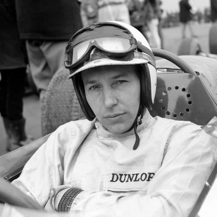 Race car driver in vintage gear, sitting in a cockpit, wearing a helmet and goggles, with a focused expression.
