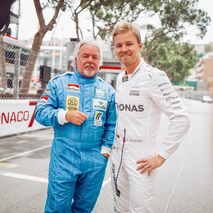 Two racing drivers in blue and white suits pose at a Monaco Grand Prix track, showcasing their team pride and camaraderie.