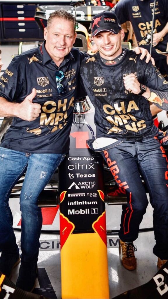 Two individuals celebrating in a racing garage, wearing Champions shirts by Red Bull Racing, next to a race car.