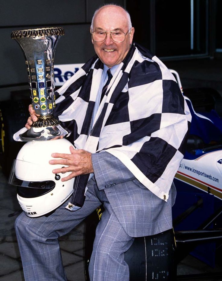 Man in checkered flag drapes holding racing trophy and helmet, sitting on tire, standing near race car.