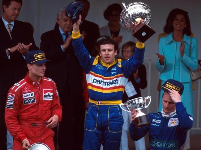 Race car driver celebrates victory on podium with trophy, flanked by two competitors. Emphasize motorsport success and celebration.