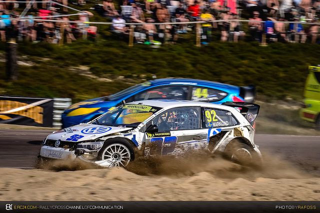 Rallycross race action with two cars speeding on a dirt track, surrounded by spectators, highlighting dynamic motorsport excitement.