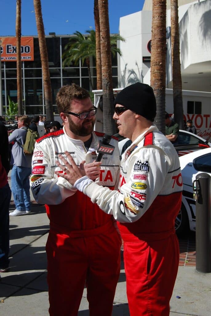 Two race car drivers in Toyota suits converse outdoors at a motorsport event, surrounded by spectators and palm trees.