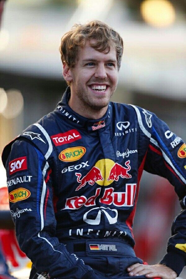 Race car driver smiling in a full Red Bull racing suit, featuring sponsor logos, showing excitement before the event.