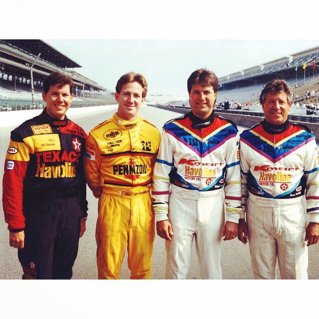 Four race car drivers in colorful uniforms standing on a racetrack, showcasing their team sponsors.