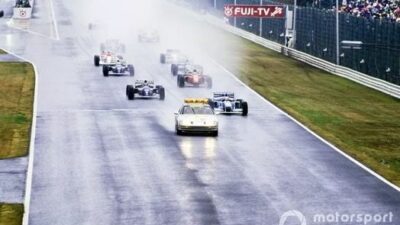 Race cars following a safety car on a wet track, creating spray at a rainy circuit with a crowd in the background.