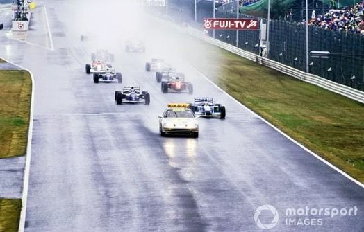 Race cars following a safety car on a wet track, creating spray at a rainy circuit with a crowd in the background.