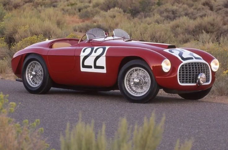 Vintage red race car with number 22, parked on a road in a scenic landscape, showcasing classic design and style.