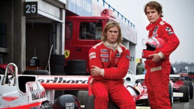 Two race car drivers in red suits pose beside classic Formula 1 cars in a pit lane, capturing a vintage racing vibe.
