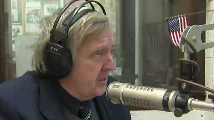 Man with headphones speaking into a microphone in a radio studio setting featuring a small American flag.
