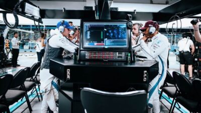 Race team strategizes in pit garage with computers and headsets during a car competition event.