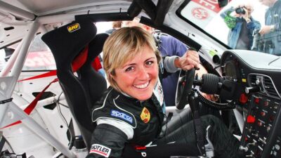 Smiling race car driver in black suit sitting inside a car, gripping the steering wheel, with photographers capturing the moment.