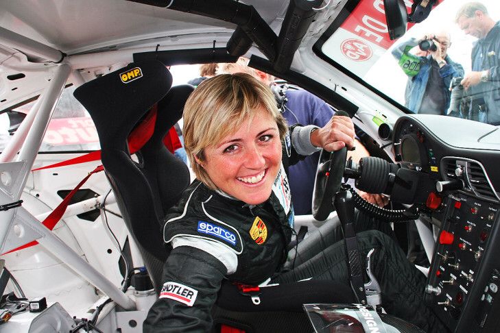 Smiling race car driver in black suit sitting inside a car, gripping the steering wheel, with photographers capturing the moment.