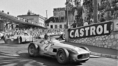 Vintage car race with classic vehicles on a street circuit, drivers navigating a turn, surrounded by spectators and Castrol signage.