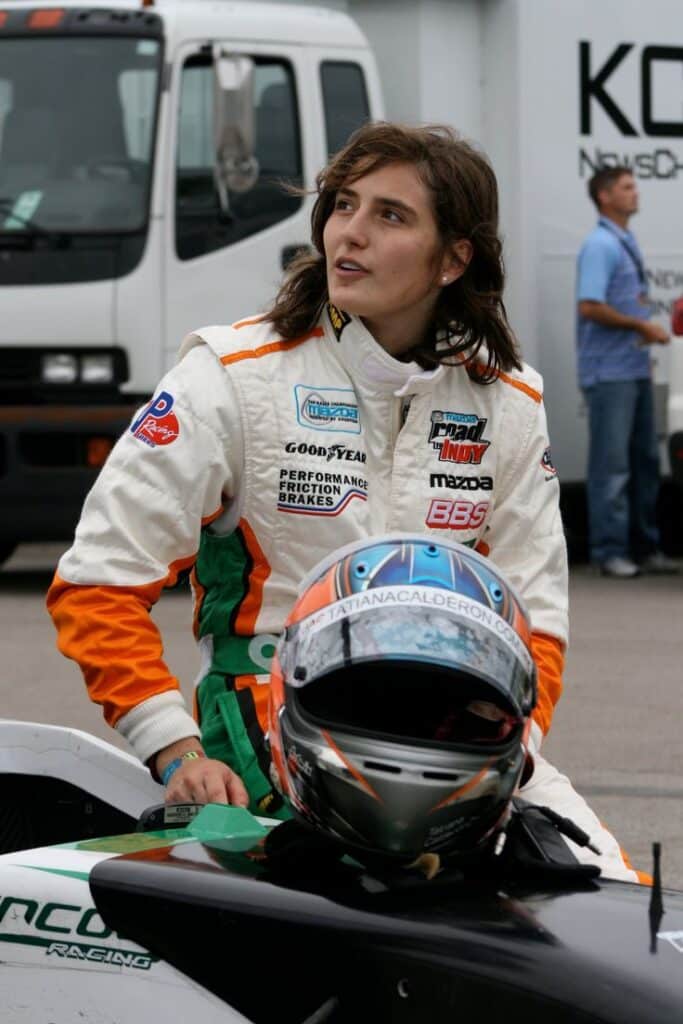 Race car driver in a suit stands by a helmet on a vehicle, preparing for a race, with a truck in the background.