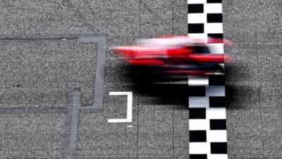 Blurred red race car crossing finish line on checkered asphalt track, showcasing speed and competition.