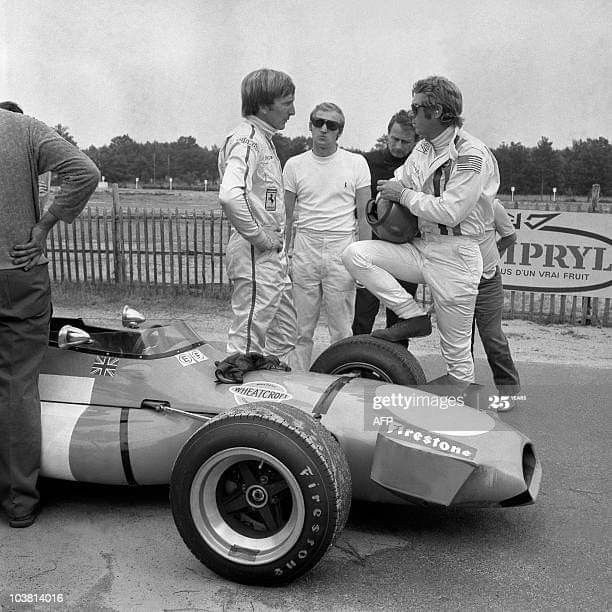 Vintage racing drivers discuss strategy beside a classic race car on a track day, showcasing teamwork and automotive history.