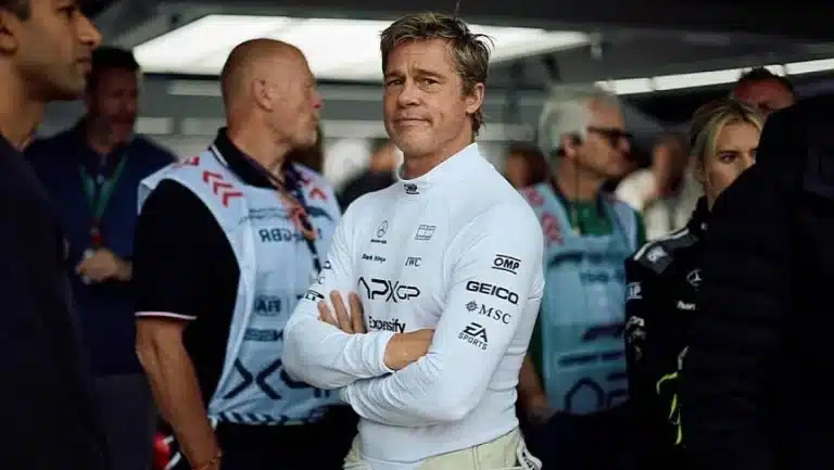Race car driver in a white suit stands confidently in a garage surrounded by team members, showcasing motorsport focus and determination.