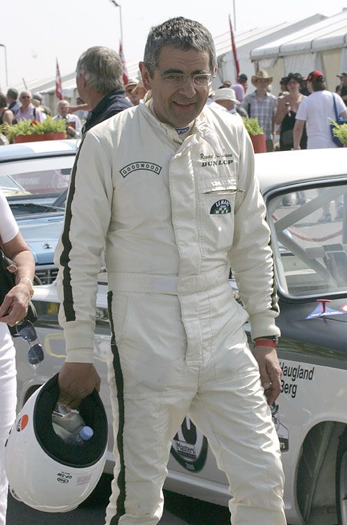 Man in racing suit holding helmet at a car event, surrounded by people and cars, on a sunny day.