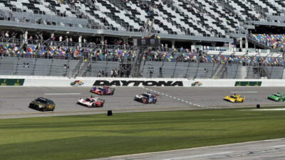 Race cars speeding on Daytona International Speedway track in front of a large crowd in the stands.