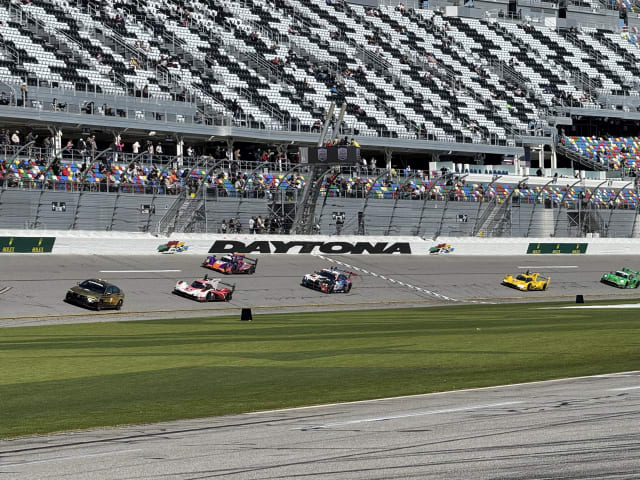 Race cars speeding on Daytona International Speedway track in front of a large crowd in the stands.