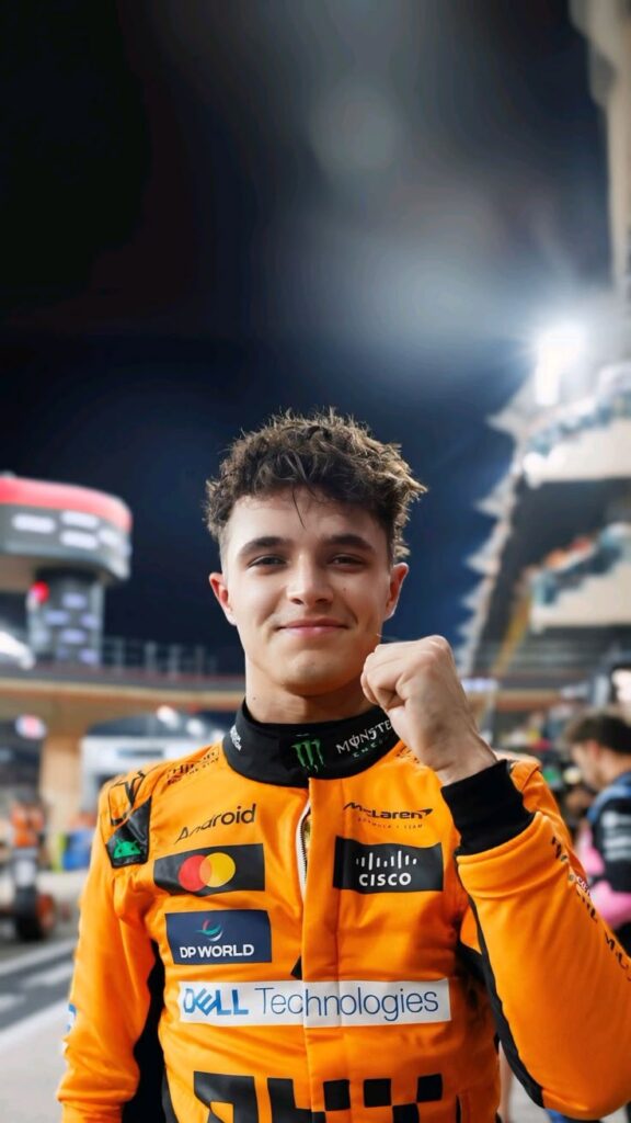 Race car driver celebrating victory, wearing orange suit with sponsor logos. Night race track background.