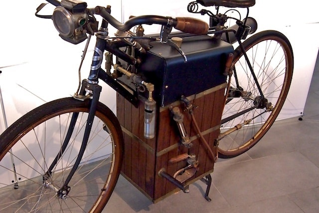 Vintage motorized bicycle with wooden and metal components displayed indoors, showcasing early engineering innovation.