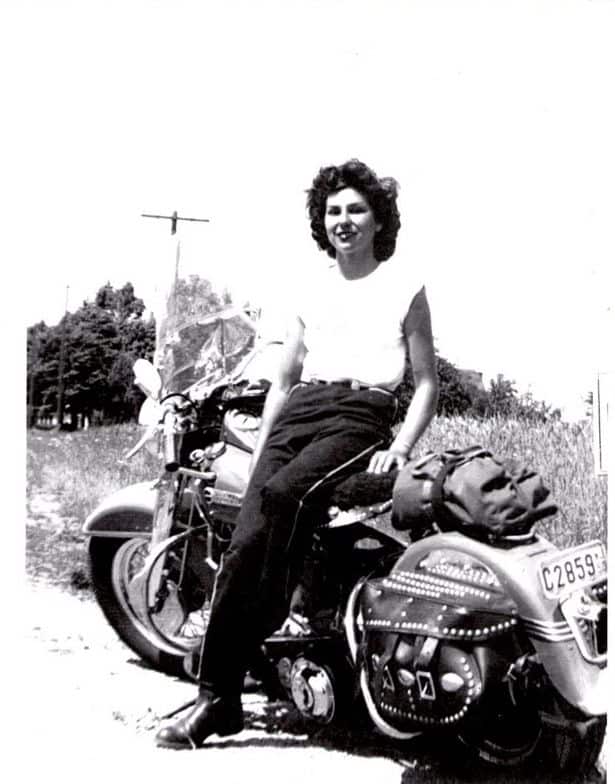 Woman sitting on a vintage motorcycle in a rural setting, 1950s style, showcasing freedom and adventure.