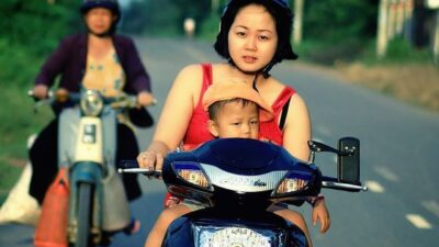 Woman and child riding a scooter on a rural road, with another person following in the background on a motorcycle.