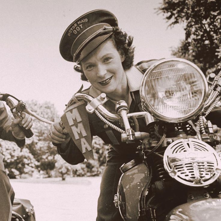 Vintage photo of a smiling woman in a Motor Maid hat, leaning on a classic motorcycle outdoors.