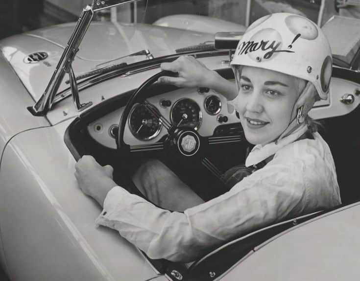 Vintage photo of a woman in a racing helmet sitting in a classic sports car, smiling at the camera. Racing nostalgia.