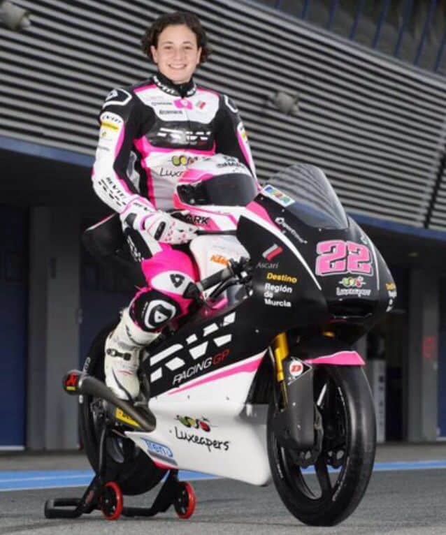 Motorcycle racer in pink and white gear poses on a branded bike with number 22 at a racing circuit.