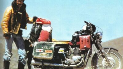 Man with retro motorcycle packed for adventure, standing in rugged landscape under blue sky.