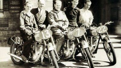 Vintage group of five men with three numbered racing motorcycles outside a brick building, showcasing retro motorsport style.